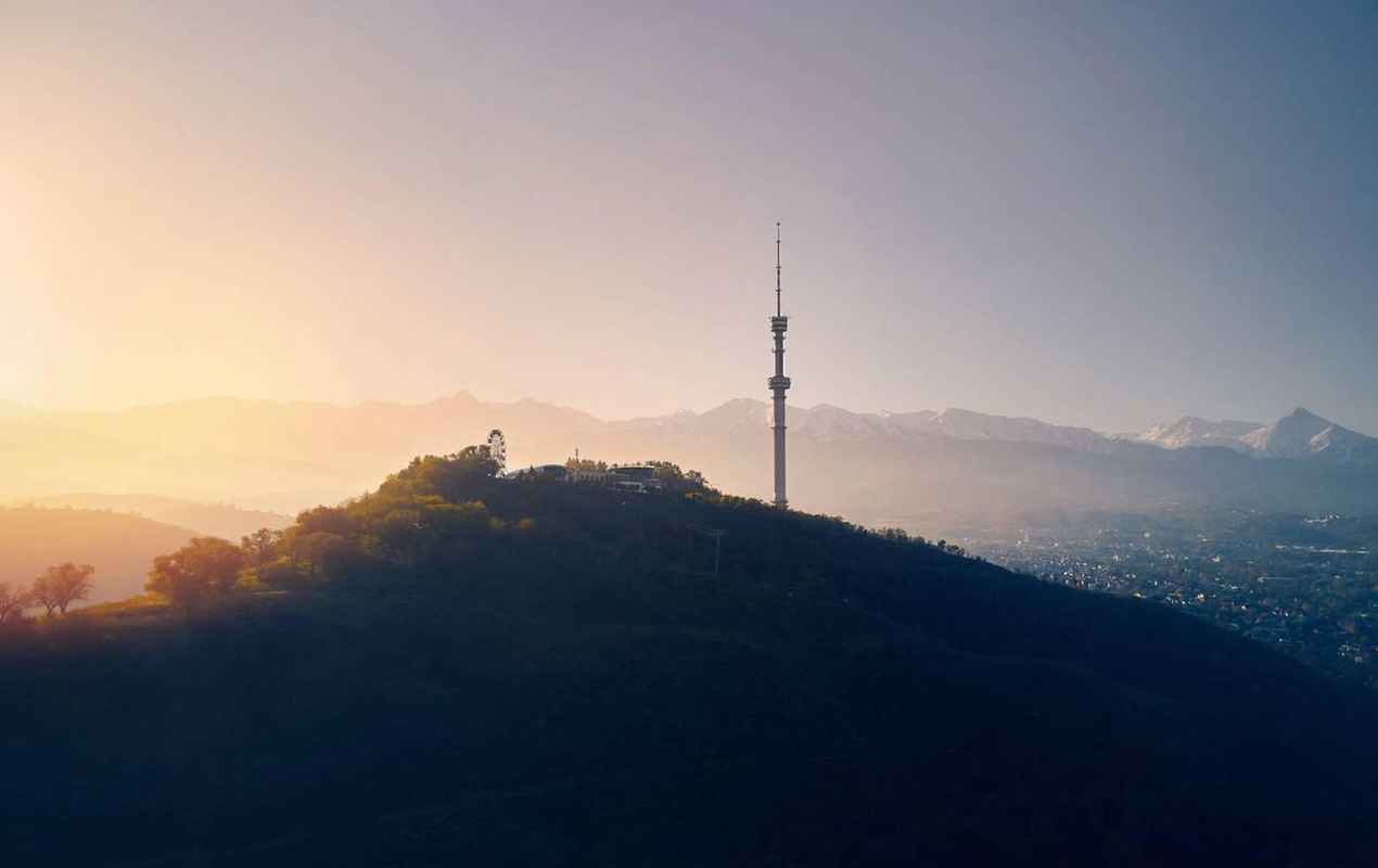 Aerial drone shot of symbol Almaty city high TV tower and park at Koktobe hill against snow mountains at sunrise in Kazakhstan